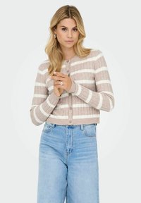 Korte cardigan in beige met witte strepen, ribbeltextuur, knoopsluiting aan de voorkant en een ronde hals. Gecombineerd met lichtblauwe high-waisted jeans.