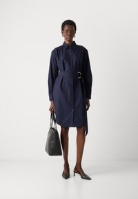 Marineblauw knielang jurkje met een ceintuur in de taille, lange mouwen en een rechte snit. Het model houdt een grijze handtas vast en draagt zwarte hakken.