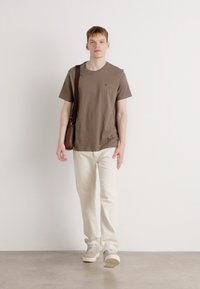 Braunes Baumwoll-T-Shirt mit kurzen Ärmeln, weiße Jeans und beigetöne Sneaker. Eine braune Leder-Umhängetasche hängt über der rechten Schulter.