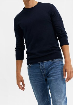 Mann trägt einen dunkelblauen Pullover mit langen Ärmeln und blaue Jeans, steht vor einem schlichten hellen Hintergrund.