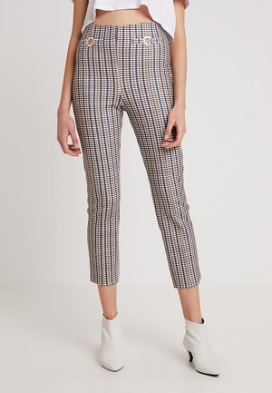 Femme portant un pantalon slim à taille haute à carreaux avec des détails de boutons dorés, associé à des bottines blanches à la cheville et un top blanc court.