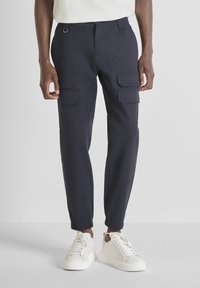 Man in een donkerblauwe cargobroek met elastische boorden en witte sneakers, staand tegen een effen lichte achtergrond.