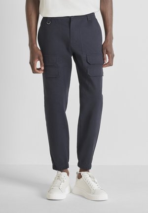 Homme portant un pantalon cargo bleu marine avec des poignets élastiques et des baskets blanches, se tenant devant un fond clair.