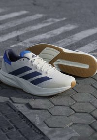 Chaussures de running blanches avec des bandes bleu marine et des accents bleus posées sur un pavement gris texturé, une chaussure montrant la semelle en gomme.