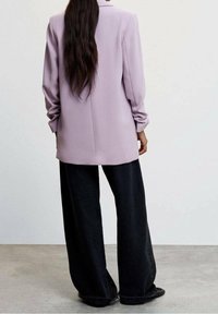 Veste légère lavande à manches longues et coupe décontractée, associée à un pantalon large noir. Tissu à la texture lisse et design minimaliste.