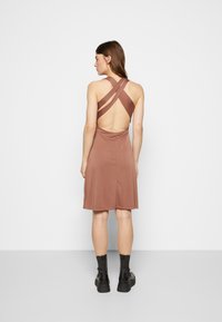 Vestido castanho, sem mangas, com alças cruzadas e costas abertas. Tecido macio, até ao joelho, com uma saia fluída, combinado com botas pretas até ao tornozelo.