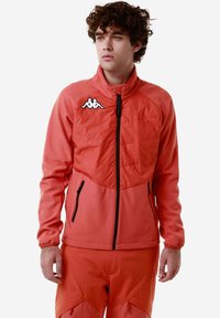 Giacca corallo in tessuto testurizzato, colletto alto e chiusura con zip. Presenta cerniere nere e logo bianco sul petto.