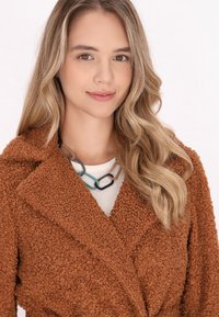 Brązowy płaszcz teddy o teksturowanej, puszystej powierzchni; z kołnierzem. Pod spodem jasny top z naszyjnikiem w wzór łańcucha.