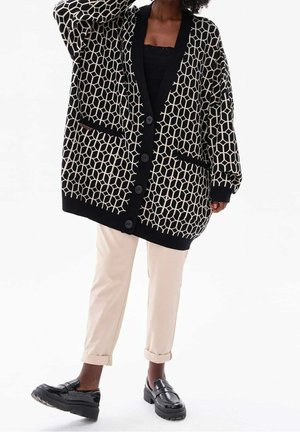 Person trägt eine schwarz-weiß geometrisch gemusterte, übergroße Strickjacke, cremefarbene Hose und schwarze Loafer und steht vor einem schlichten Hintergrund.