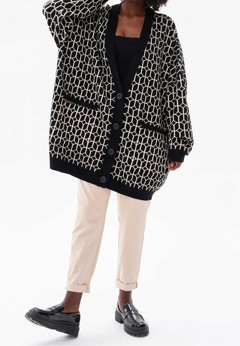 Personne portant un cardigan oversize à motifs géométriques noirs et blancs, un pantalon crème et des mocassins noirs, debout devant un fond uni.