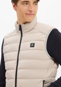 Junger Mann trägt eine beigen gesteppte Weste mit MOP-Logo über einem schwarzen gerippten Pullover, schaut zur Seite mit einem leichten Lächeln.