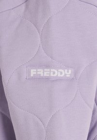 Tela acolchada color lavanda con un patrón de curvas. Una etiqueta blanca con la palabra "FREDDY" está cosida en el material. Textura suave.