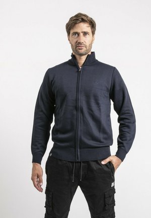 Homme debout avec une expression sérieuse, portant un sweat à fermeture éclair bleu marine et un pantalon cargo noir, sur un fond clair uni.