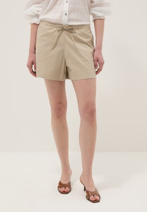 Frau trägt beige Shorts mit Kordelzug, weiße Bluse mit Knöpfen und Puffärmeln sowie braune offene Sandalen mit Absatz, steht vor einfarbigem Hintergrund.