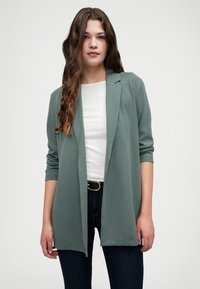 Grønn drapert langermet cardigan laget av mykt stoff, med åpen front og løs passform, kombinert med en hvit topp og mørke jeans.