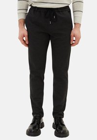 Černé teplákové kalhoty z měkké látky s elastickým pasem se šňůrkou a zúženými nohavicemi, doplněné černými botami.