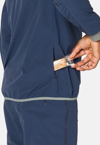 Marineblauer Windbreaker mit einer Reißverschlusstasche. Der entspannte Schnitt hat einen kontrastierenden grünen Rand und ein Objekt wird aus der Seitentasche entnommen.