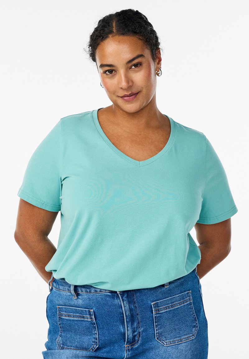 Frau mit lockigen Haaren, die Creolen, ein hellblaues V-Ausschnitt-T-Shirt und eine hochgeschnittene blaue Jeans mit Fronttaschen trägt, posiert mit den Händen hinter dem Rücken.