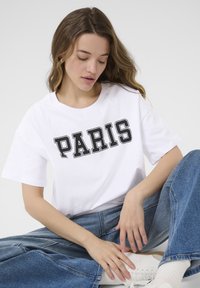T-shirt blanc à manches courtes avec un texte noir en gras, "PARIS". Assorti à un jean large en denim bleu et des baskets blanches avec des accents beiges.