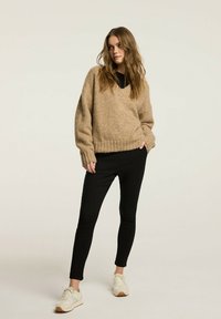 Beżowy sweter z dekoltem w serek, w oversize'owym kroju, z prążkowanym wykończeniem, noszony na czarnej bluzce, w zestawieniu z wąskimi czarnymi spodniami i białymi sneakersami. Gładka faktura.