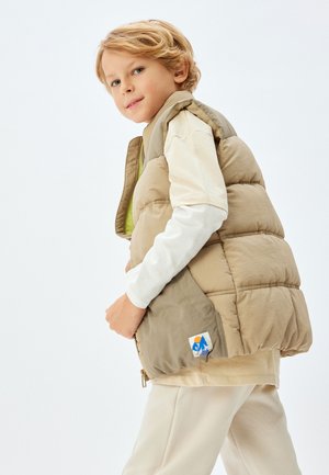 Chaleco puffer beige con cuello alto y bolsillos laterales, que presenta un logo de montaña azul. Lleva una camisa de manga larga ligera debajo.