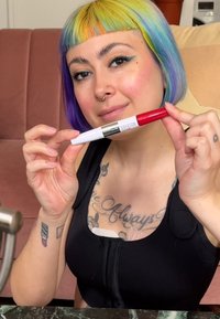 Marker do ust z białą obudową i czerwoną nakrętką, oznaczony napisem "Super Stay". Osoba trzyma go na tle różowej kanapy.