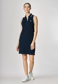 Vestito polo senza maniche di colore navy, realizzato in cotone, con colletto con dettagli in azzurro chiaro e un piccolo logo sul petto, abbinato a sneakers bianche.
