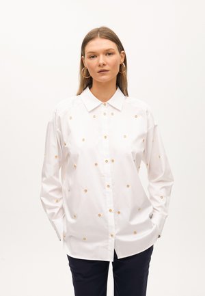 White button-up shirt with gold circular embellishments, long sleeves, and a classic collar. Worn over navy pants.