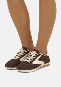 Scarpe sportive marroni con dettagli bianchi, in materiale scamosciato e sintetico, punta arrotondata e suola piatta in gomma. I lacci sono beige.