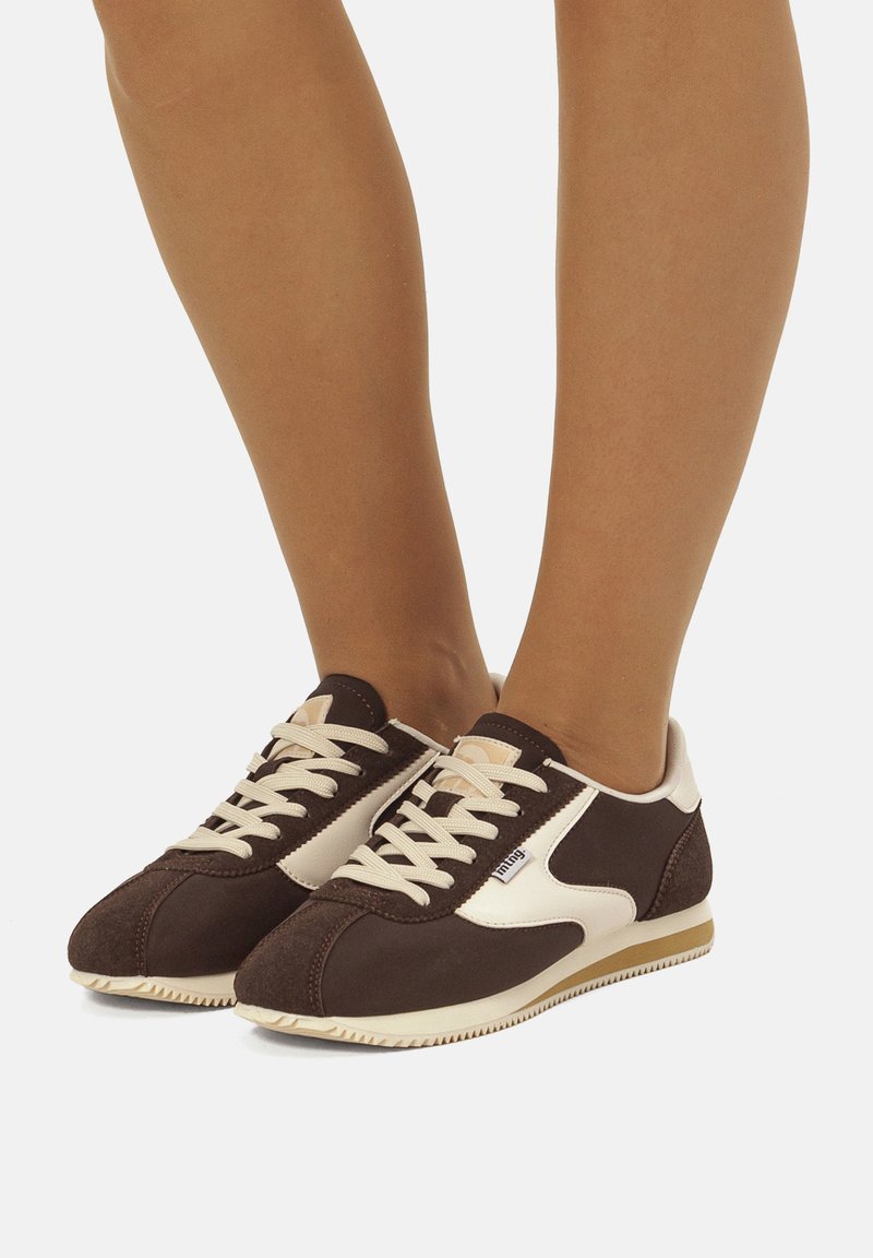 Scarpe sportive marroni con dettagli bianchi, in materiale scamosciato e sintetico, punta arrotondata e suola piatta in gomma. I lacci sono beige.