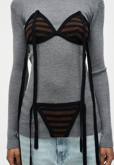 Sous-vêtements rayés marron et noirs portés par-dessus un pull gris côtelé avec un jean en denim bleu clair, avec de longues bretelles noires pendantes.