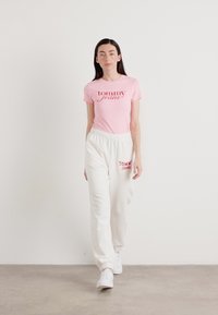 Roze katoenen T-shirt met het "tommy jeans" logo in het rood, gecombineerd met witte sweatpants met hetzelfde logo. Model draagt witte sneakers.