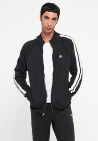 Black zip-up jacket with white side stripes, ribbed collar, and a small embroidered logo on the chest. Model poses with hands in pockets.