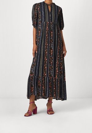 Robe maxi en navy avec des rayures verticales, des motifs floraux, des manches courtes bouffantes, un décolleté en V avec un nœud, et un ourlet volanté. Portée avec des sandales à talons roses.