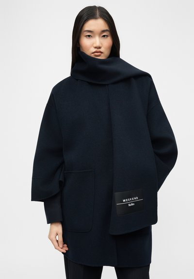 Γυναίκα με μακριά μαύρα μαλλιά φοράει μαύρο, φαρδύ παλτό και ταιριαστό κασκόλ με την ετικέτα "WEEKEND MaxMara" μπροστά σε απλό γκρί φόντο.