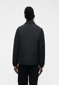 Homem com dreadlocks, vestindo um casaco preto e calças pretas, está de costas contra um fundo branco simples.