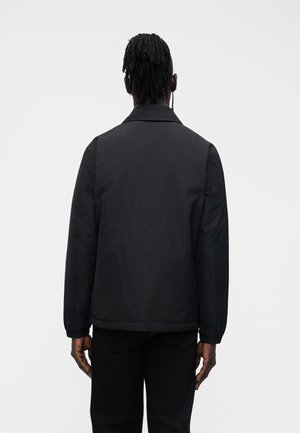 Mees dredsidega, seljas must jakk ja mustad püksid, seisab valge taustaga vastu paljut musta tausta seljaga.