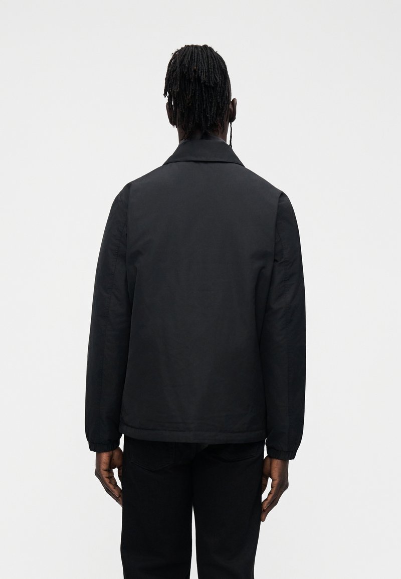 Homem com dreadlocks, vestindo um casaco preto e calças pretas, está de costas contra um fundo branco simples.