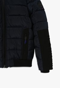 Mörkblå vadderad jacka med svarta armbågsfläckar, dragkedjeficka med blå dragflik och ribbad nederkant och ärmslut.