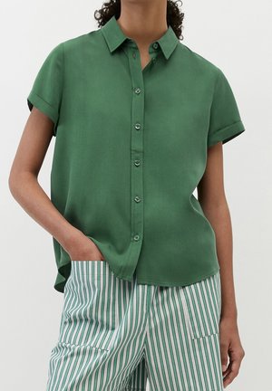 Persoon draagt een groen knoopoverhemd met korte mouwen, gecombineerd met groen-wit verticaal gestreepte broek, één hand in de zak.
