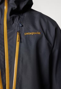Jachetă Patagonia de culoare bleu închis, cu fermoare galbene strălucitoare, glugă ajustabilă și logo brodat pe piept. Material neted, rezistent la apă.