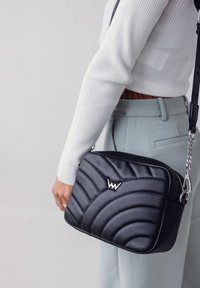 Prošívaná černá kožená taška přes rameno se stříbrným logem, plochý design a nastavitelný popruh, s lesklým zipem a texturovanými akcenty.