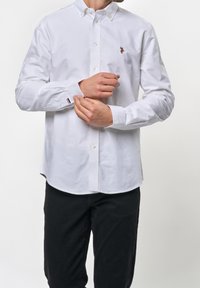 Camicia bianca in cotone con colletto a pieghe, maniche lunghe e piccolo logo ricamato sul petto.
