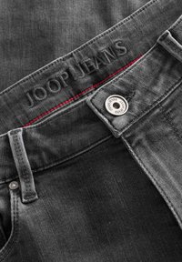 Tummanharmaat farkut, joissa on JOOP! JEANS -logo vyötäröllä ja punaiset ompeleet. Metallinappi ja standardi viiden taskun muotoilu.
