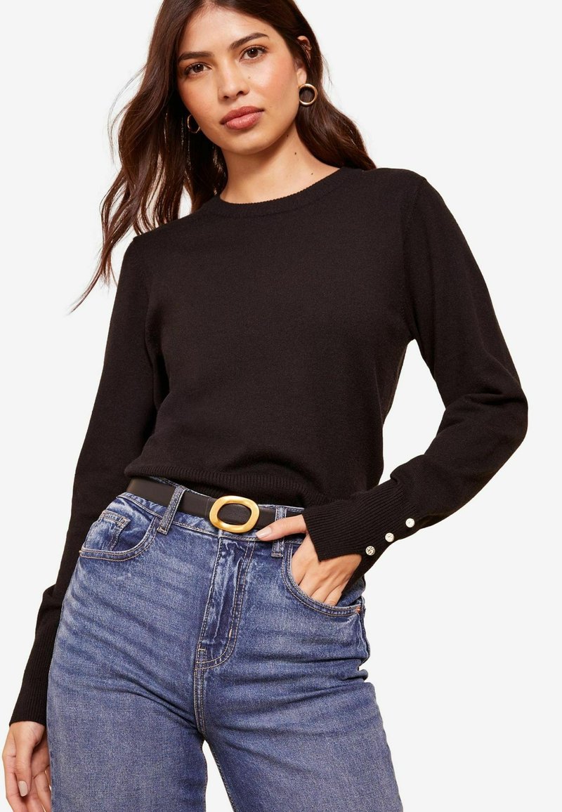 Un pulover negru tricotat cu manșete cu deraiere, dotat cu accente de nasturi, asortat cu blugi denim cu talie înaltă și o curea neagră cu o cataramă rotundă aurie.