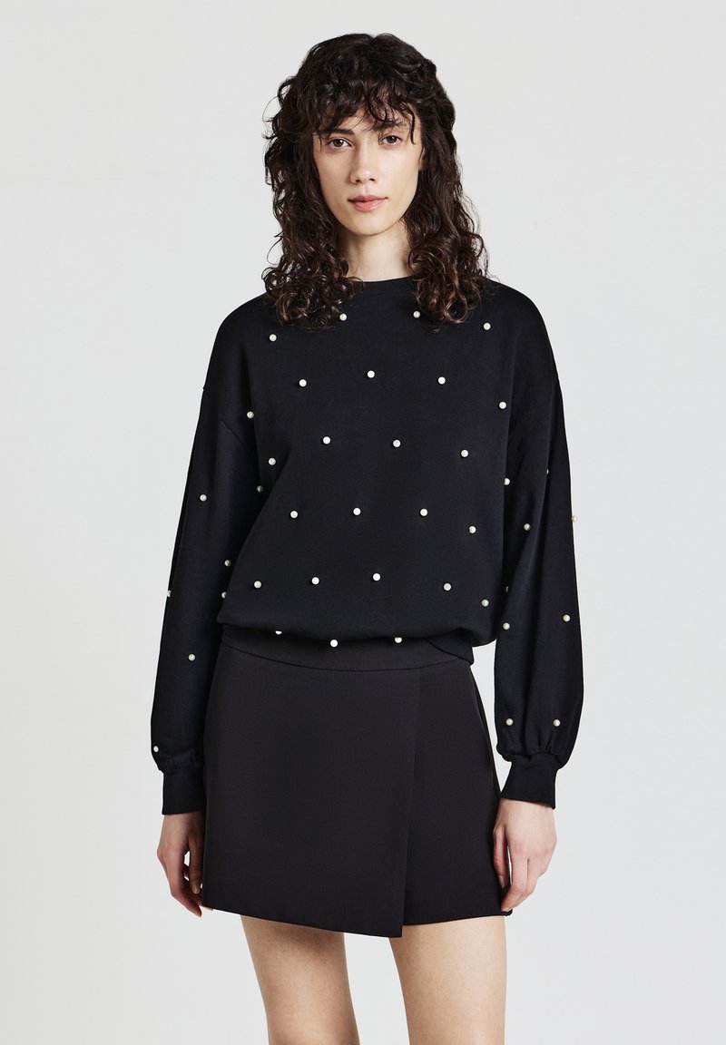 Felpa nera a maniche lunghe con decorazioni di perle bianche, abbinata a una gonna nera a portafoglio. Tessuto morbido, vestibilità comoda e spalle scese.