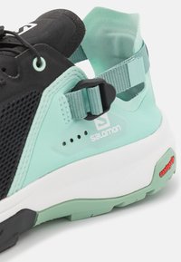 Zapatilla Salomon con parte superior de malla negra, acentos en verde menta, correa ajustable y suela blanca con el logo roja de Contagrip.