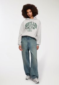 Felpa grigia con grafica "Portland" in verde e jeans in denim azzurro chiaro a vestibilità ampia. Il modello indossa scarpe da ginnastica argentate.
