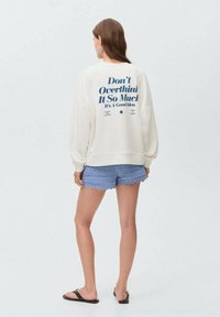 White sweatshirt with blue text on the back, paired with light blue scalloped shorts. Model wearing black sandals, standing against a light background.