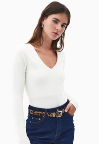 Joven mujer con cabello largo castaño, vistiendo una blusa blanca de escote en V, vaqueros azules y un cinturón de leopardo con hebilla dorada, de pie frente a un fondo blanco.